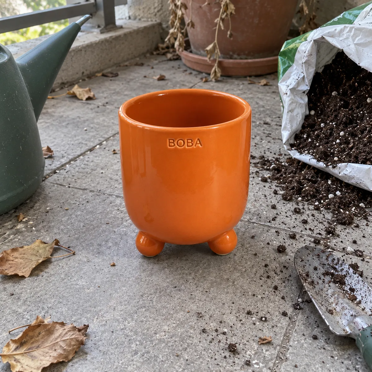 An orange footed planter on a balcony floor with spilled soil, dry leaves and a trowel