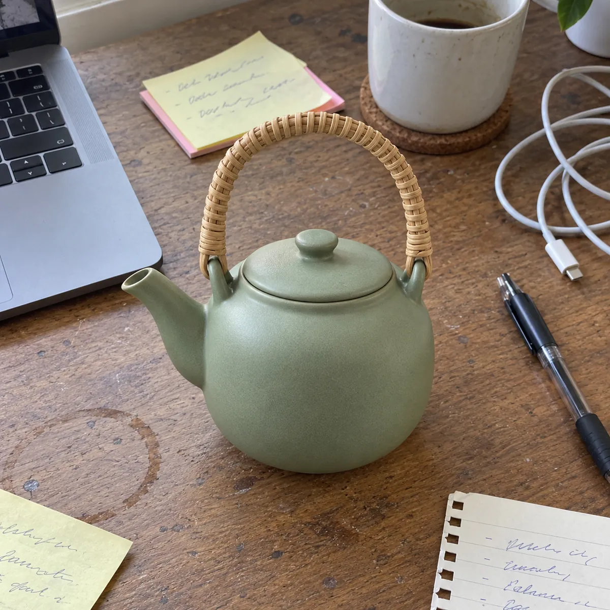 The teapot on a cluttered wooden desk next to a laptop, sticky notes and a pen