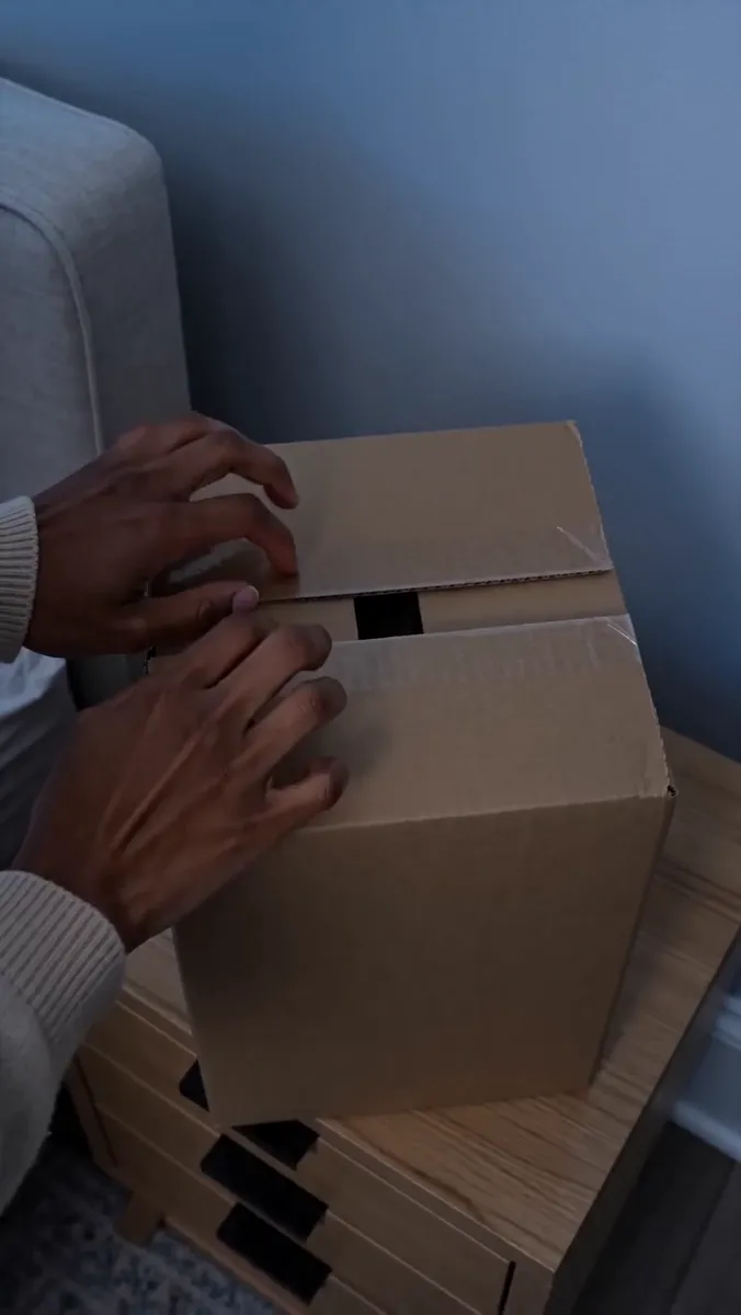 Two hands resting on a closed kraft box on a bedside table in dim light