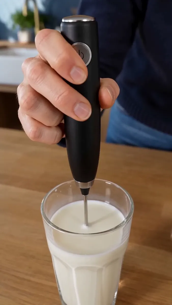 A hand holds the frother with its whisk head just under the surface of the milk